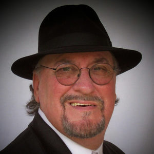White man with mustache and short beard and glasses smiling in hat button-down white shirt and black suit jacket