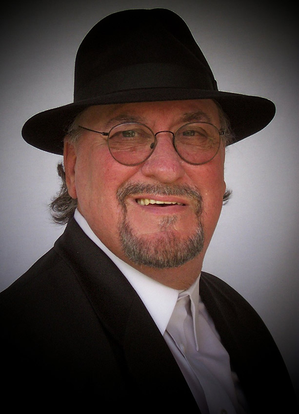 White man with mustache and short beard and glasses smiling in hat button-down white shirt and black suit jacket