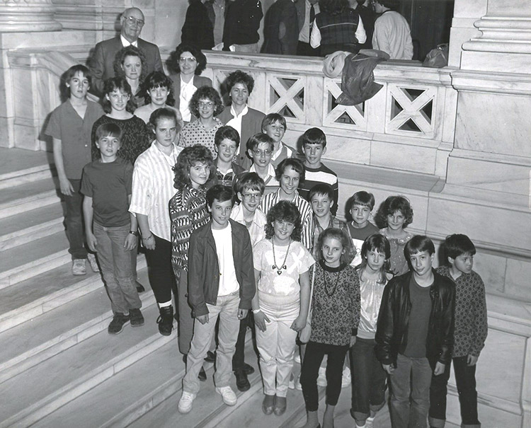 Bald white man with glasses on stone staircase with group of white children