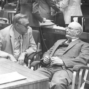 Two old white men in suits and glasses sitting speaking to each other in house chamber with men standing behind them