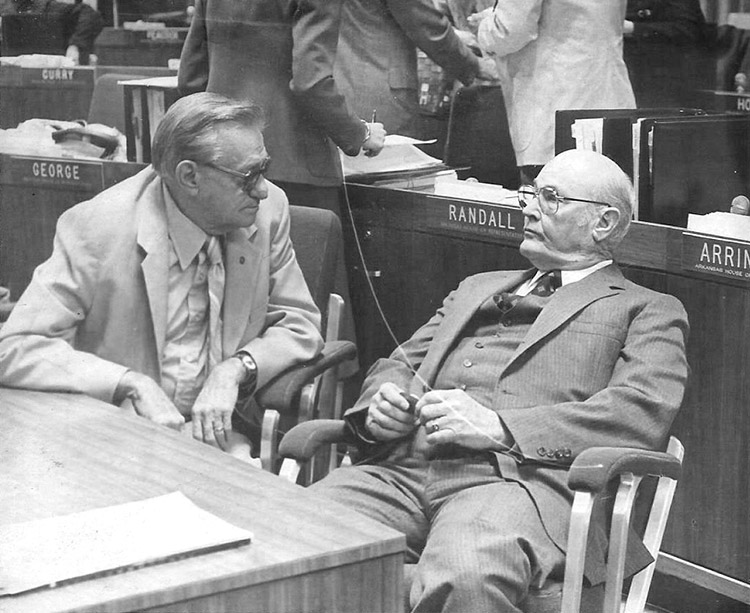 Two old white men in suits and glasses sitting speaking to each other in house chamber with men standing behind them