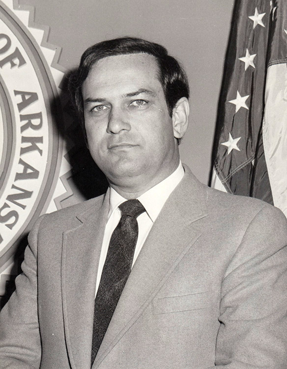 White man in suit standing with State Seal and flag behind him