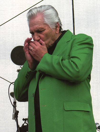 White man with gray hair in green jacket and black shirt playing harmonica