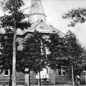 Multiple story brick building with cupola on top and trees in front yard and people sitting on front steps