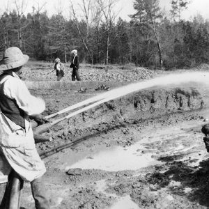 White man in hat and overalls with spraying the ground with two hoses with a man and woman in the background