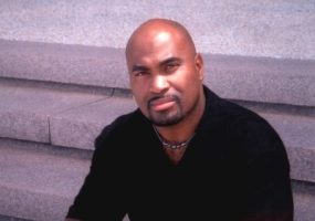 Bald African-American man with close-cropped beard and mustache sitting on steps in black shirt