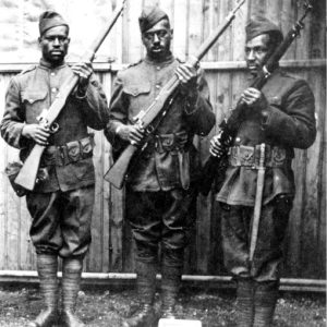 Three black soldiers uniformed holding rifles in safety position standing outside wood frame wall