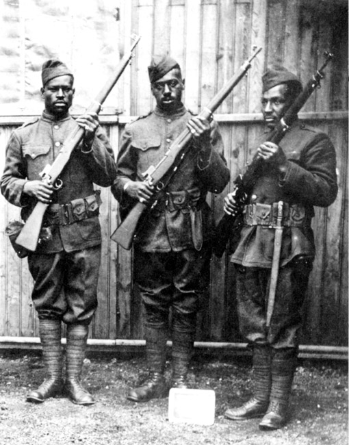 Three black soldiers uniformed holding rifles in safety position standing outside wood frame wall