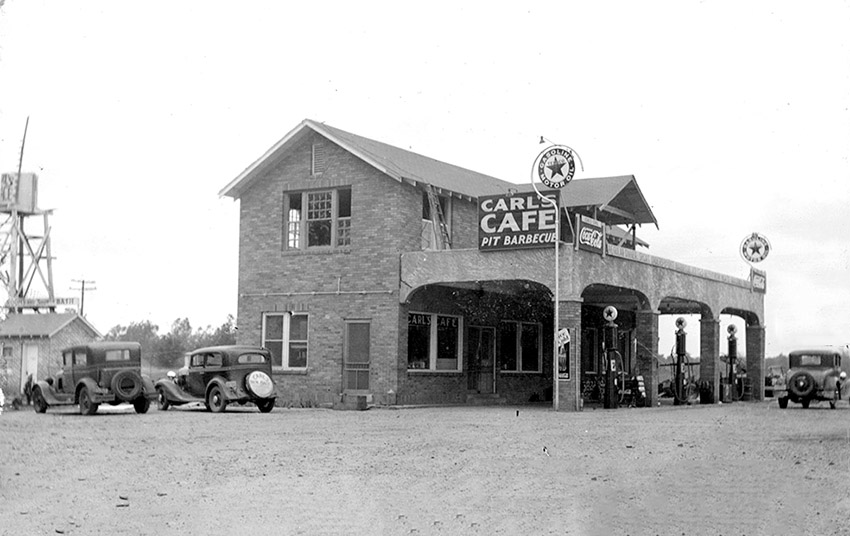 Two cars parked beside two-story restaurant building called "Carl's Cafe" with gas pumps under a canopy and car beside them