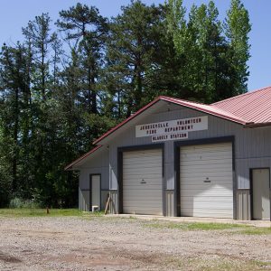 Single-story fire station building with three garage bay doors and metal siding on gravel