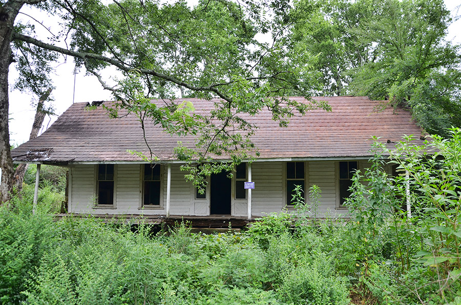 Abandoned single-story house with angled roof and covered porch in overgrown yard with trees