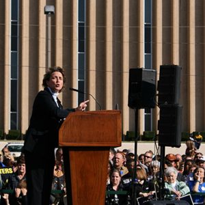 White woman in suit speaking at a lectern with crowd and building in the background