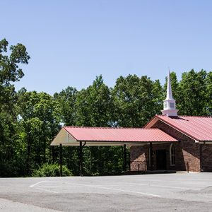 Single-story church building with steeple and covered walkway on parking lot with sign by the road