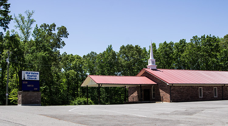 Single-story church building with steeple and covered walkway on parking lot with sign by the road