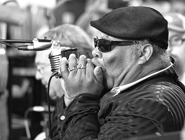 African-American man in hat and black sunglasses wearing rings playing harmonica into a microphone