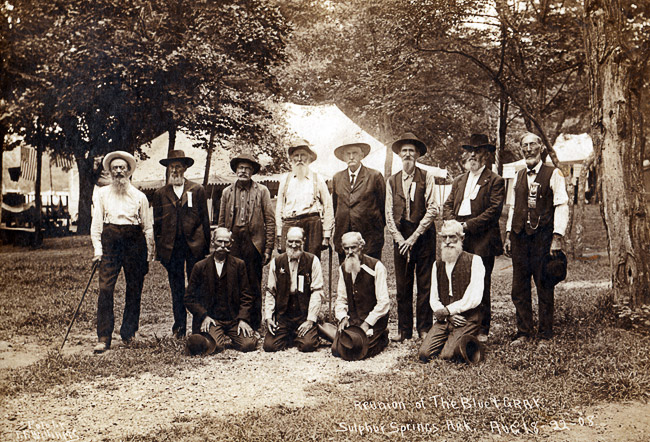 Group of sitting and standing white men mostly with beards pose for a photo with a tent in the background