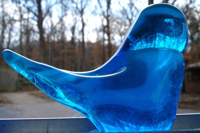 Close-up hand-blown glass bluebird simple form on metal shelf forest road and mountain in background