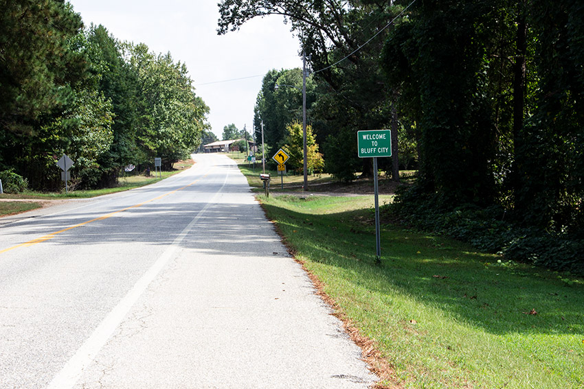 Two-lane road with green "Welcome to Bluff City" sign and yellow road sign on its right side
