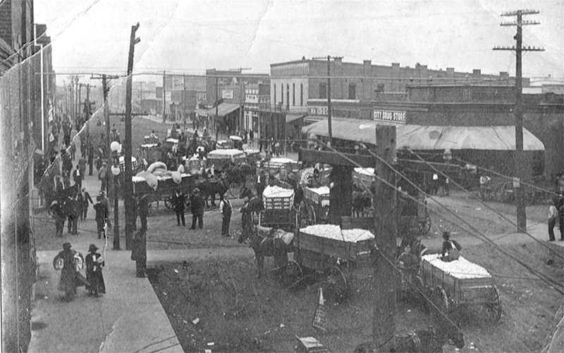 horse drawn wagons and pedestrians crowding busy street with brick buildings on either side