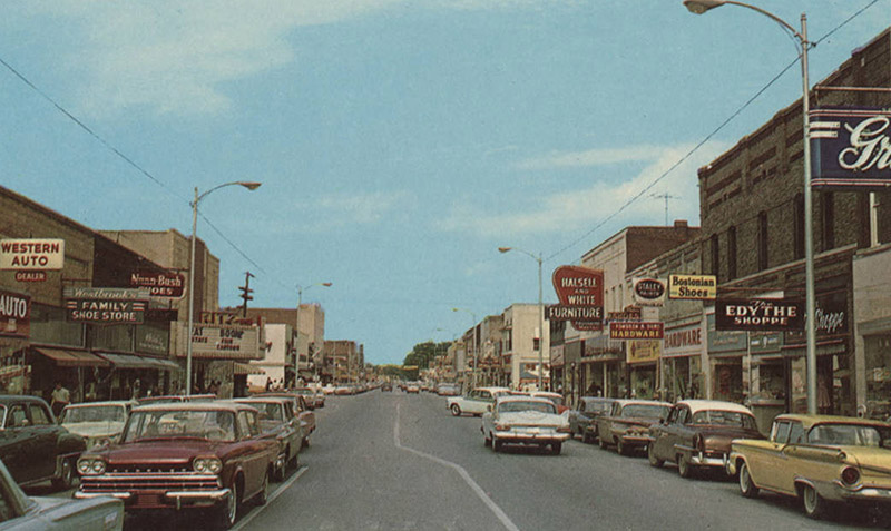 Multistory brick storefronts with signs and traffic on two-lane road lined with cars