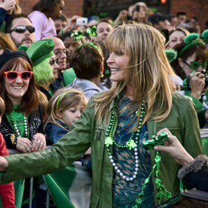 White woman in green jacket and Mardi Gras beads greeting a mixed crowd of people