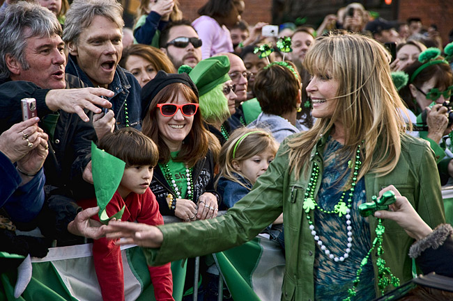 White woman in green jacket and Mardi Gras beads greeting a mixed crowd of people