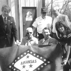 White man in suit with older white men and white woman holding up an Arkansas flag outdoors