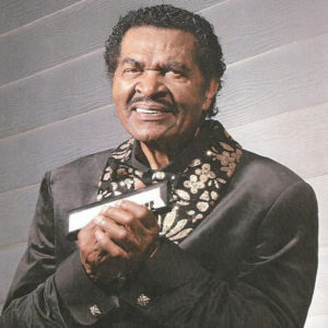 Older African-American man with mustache smiling in suit with bow tie holding a harmonica