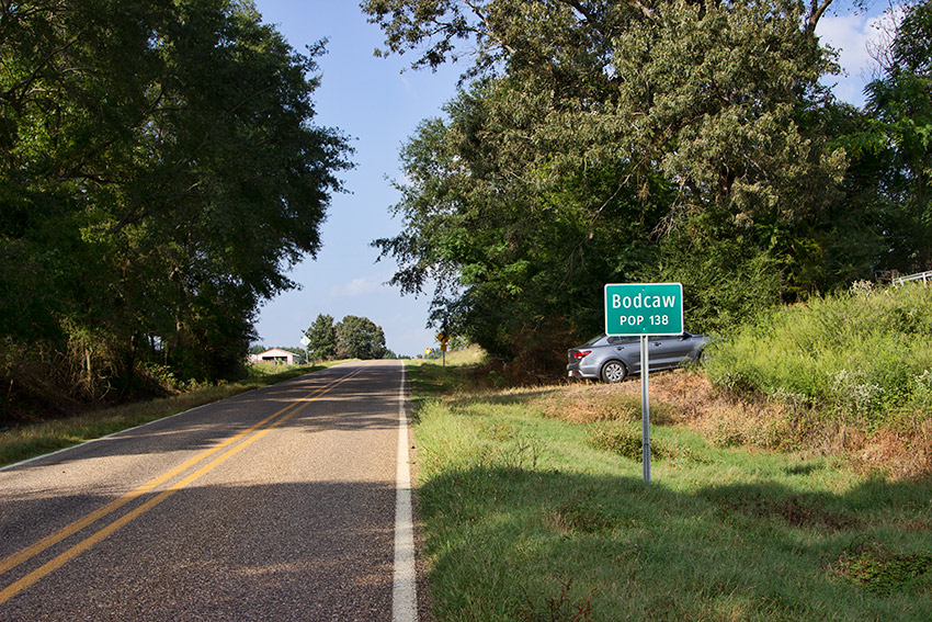 Two-lane rural road with green "Bodcaw" sign on the right side and gray car parked in driveway behind it