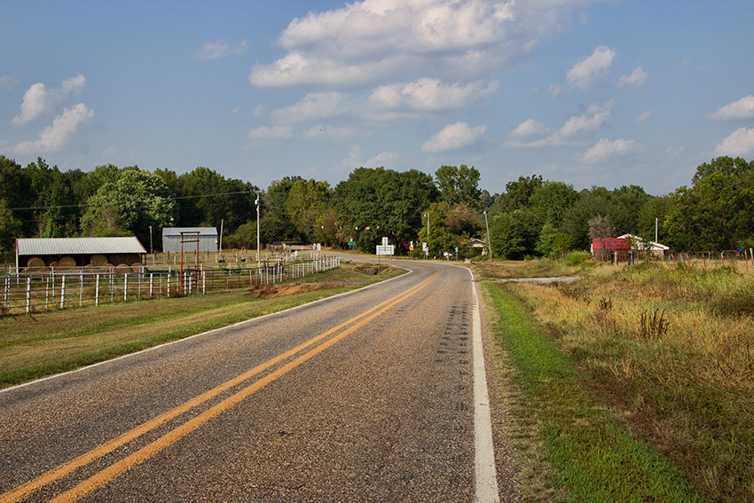 Barn and fenced-in farmland on left side of two-lane road and single-story house in field on its right side