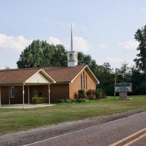Single-story brick church building with steeple next to covered concrete platform with sign on two-lane road