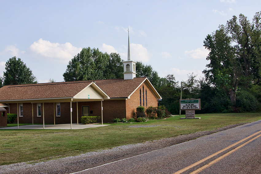 Single-story brick church building with steeple next to covered concrete platform with sign on two-lane road