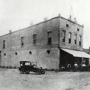 Group of men and cars outside two-story brick storefront next to single-story buildings on dirt road