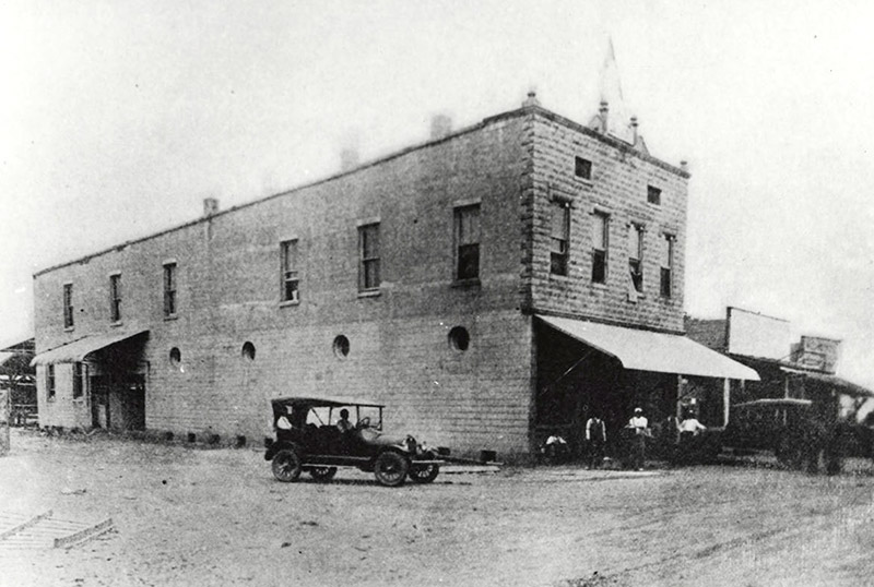 Group of men and cars outside two-story brick storefront next to single-story buildings on dirt road