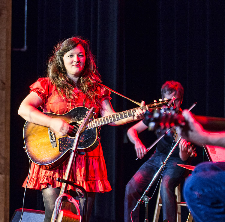 White woman in red dress playing acoustic guitar on stage