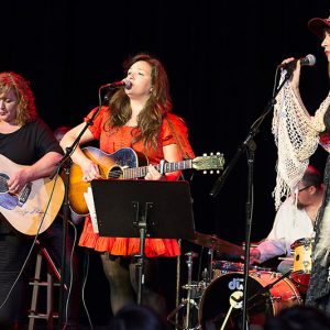 Three white women playing acoustic guitar and singing and white man on drums on stage