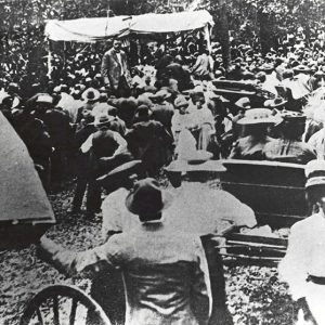 Large outdoor crowd of African-American men and women seated gathered around African-American man standing up