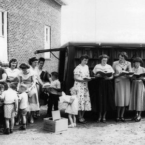 Group of white women and children gathered outside brick building looking at books from mobile library truck