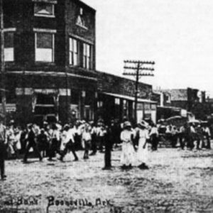 Three-story brick building and crowd on busy dirt road