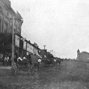 Horse drawn wagons on street with brick building on right side and multistory building with tower in the background