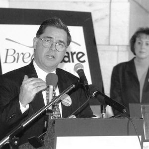 White man with glasses speaking at lectern with African-American woman with glasses and white woman standing behind him