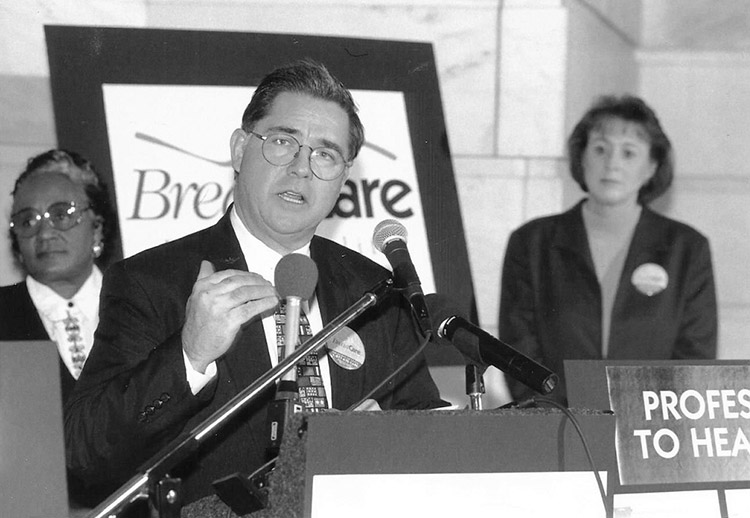 White man with glasses speaking at lectern with African-American woman with glasses and white woman standing behind him