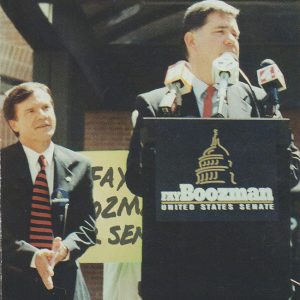 White man in suit speaking at lectern with white man in suit standing behind him