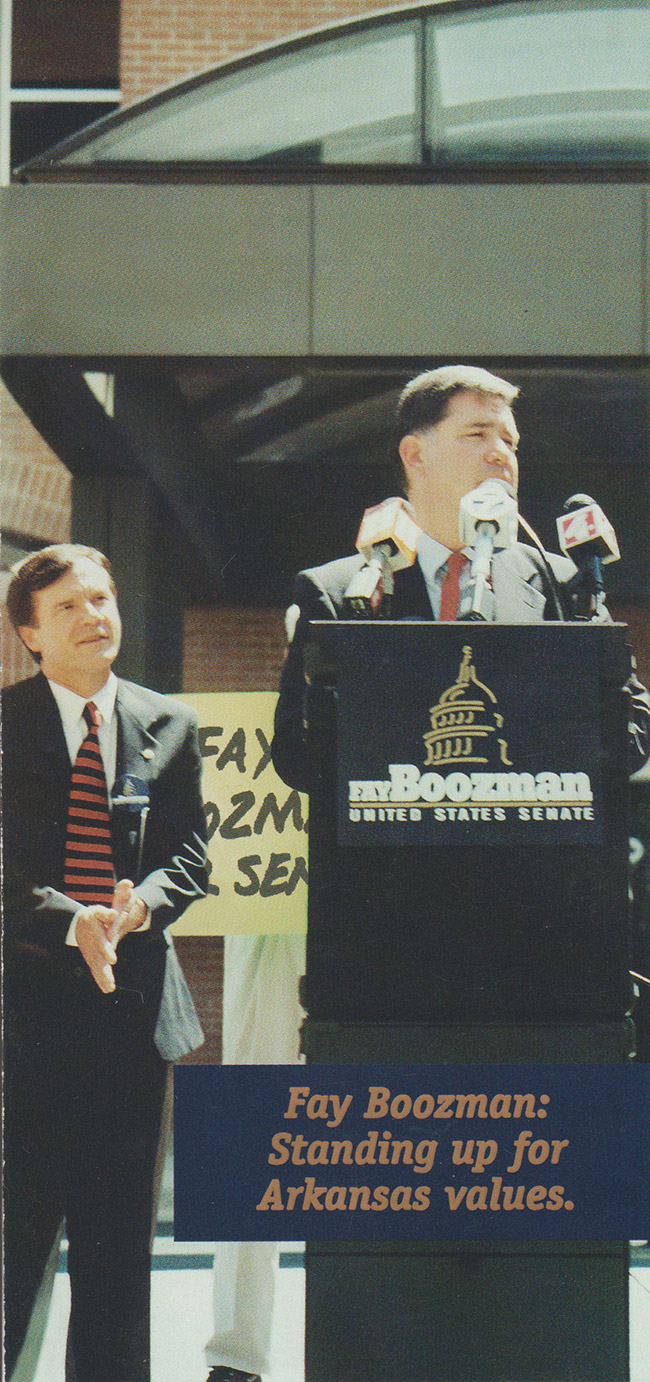 White man in suit speaking at lectern with white man in suit standing behind him