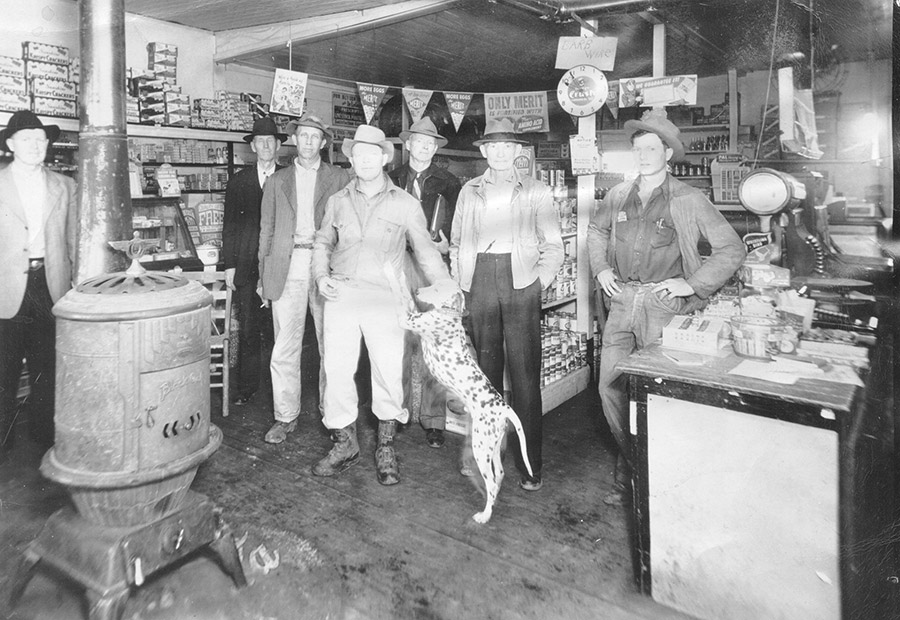 Group of white men wearing hats inside a store with a cast iron stove and Dalmatian in the foreground
