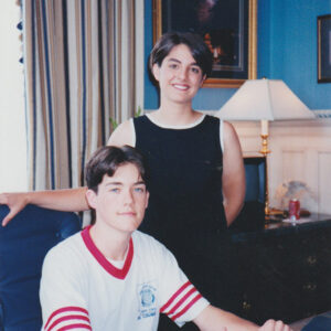 Young white woman in sleeveless dress standing with young white man sitting at desk in office
