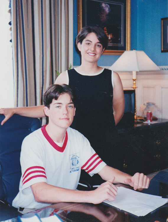 Young white woman in sleeveless dress standing with young white man sitting at desk in office