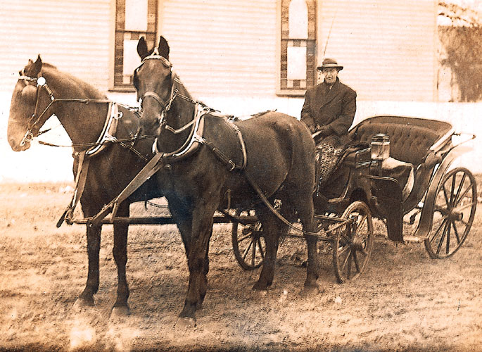 White man wearing a hat and suit sitting in a horse drawn buggy