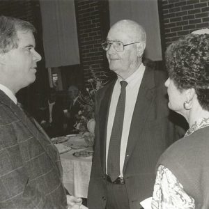 White man in suit talking to older white man with glasses in suit and older woman