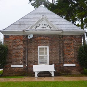 Single-story brick "County Clerk 1890" building with bench on sidewalk